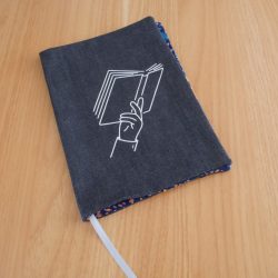 Couvre-livre fait main en jean brut avec sérigraphie blanche représentant une main tenant un livre ouvert, liseré wax coloré sur les bords