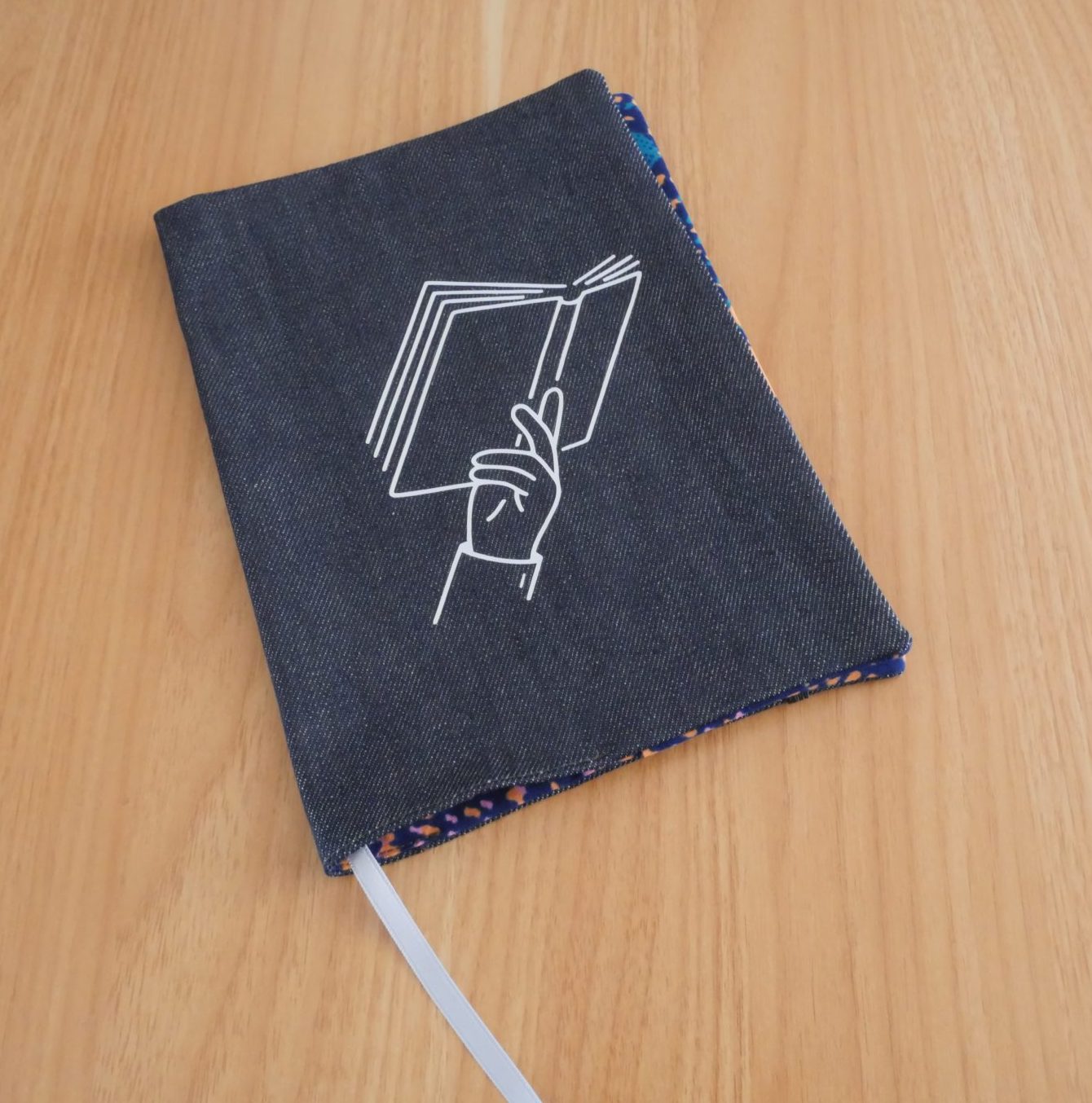 Couvre-livre fait main en jean brut avec sérigraphie blanche représentant une main tenant un livre ouvert, liseré wax coloré sur les bords