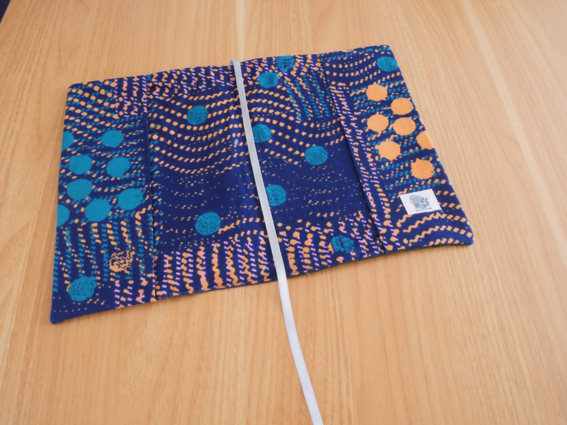 Intérieur du couvre-livre ratonetpanda ouvert, doublure wax africain bleu nuit à pois turquoise et ondulations orange, avec poche et étiquette créateur