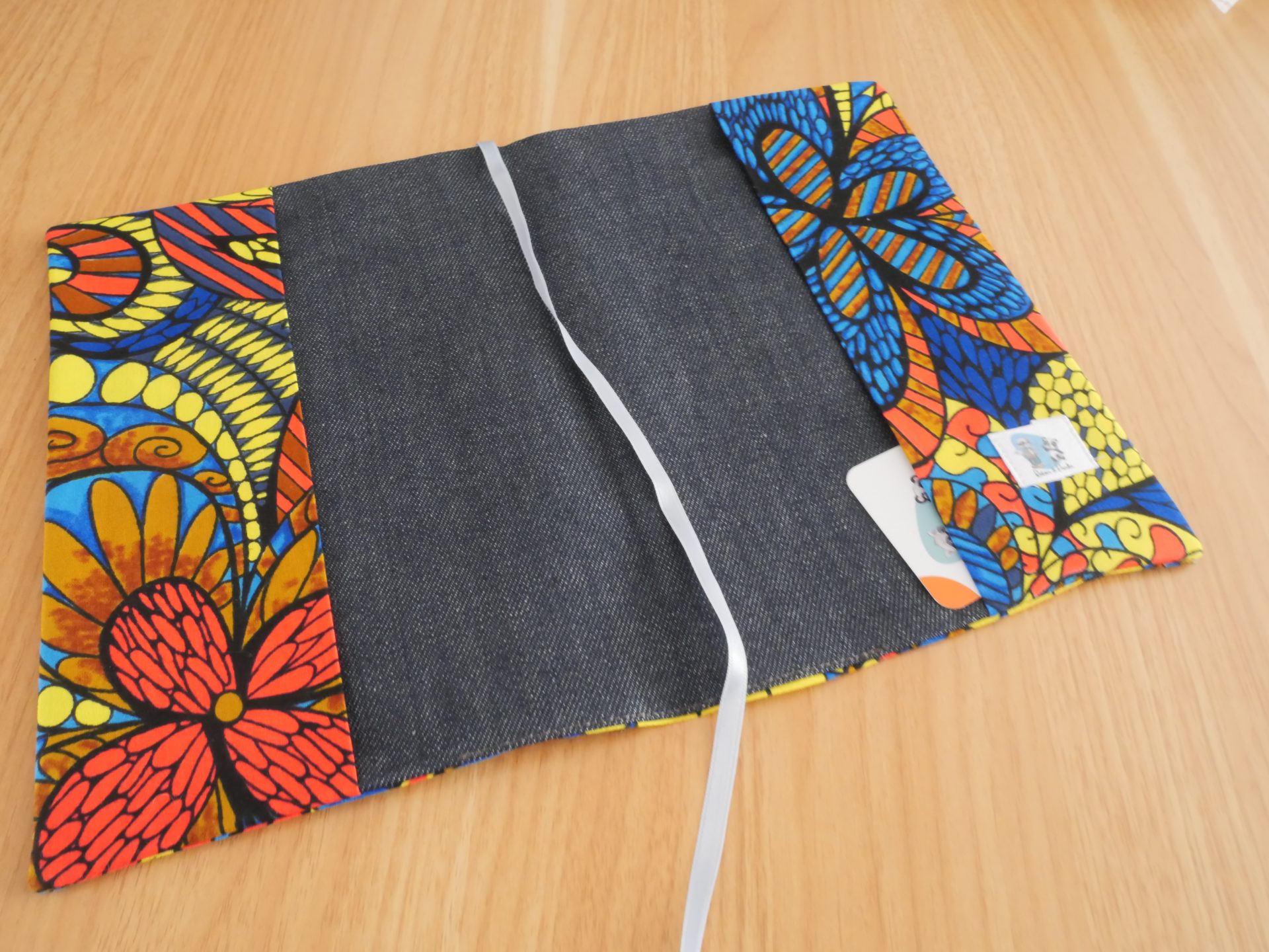 Couvre-livre fait main en tissu wax africain coloré, ouvert, avec doublure jean gris, marque-page satin blanc et poche intérieure